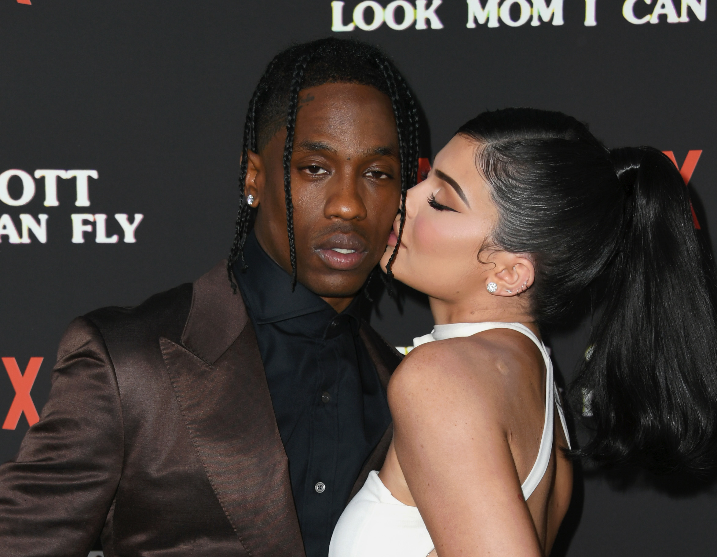 Travis Scott in a brown suit, and Kylie Jenner in a white dress, share an intimate moment on the red carpet. Kylie kisses Travis on the cheek