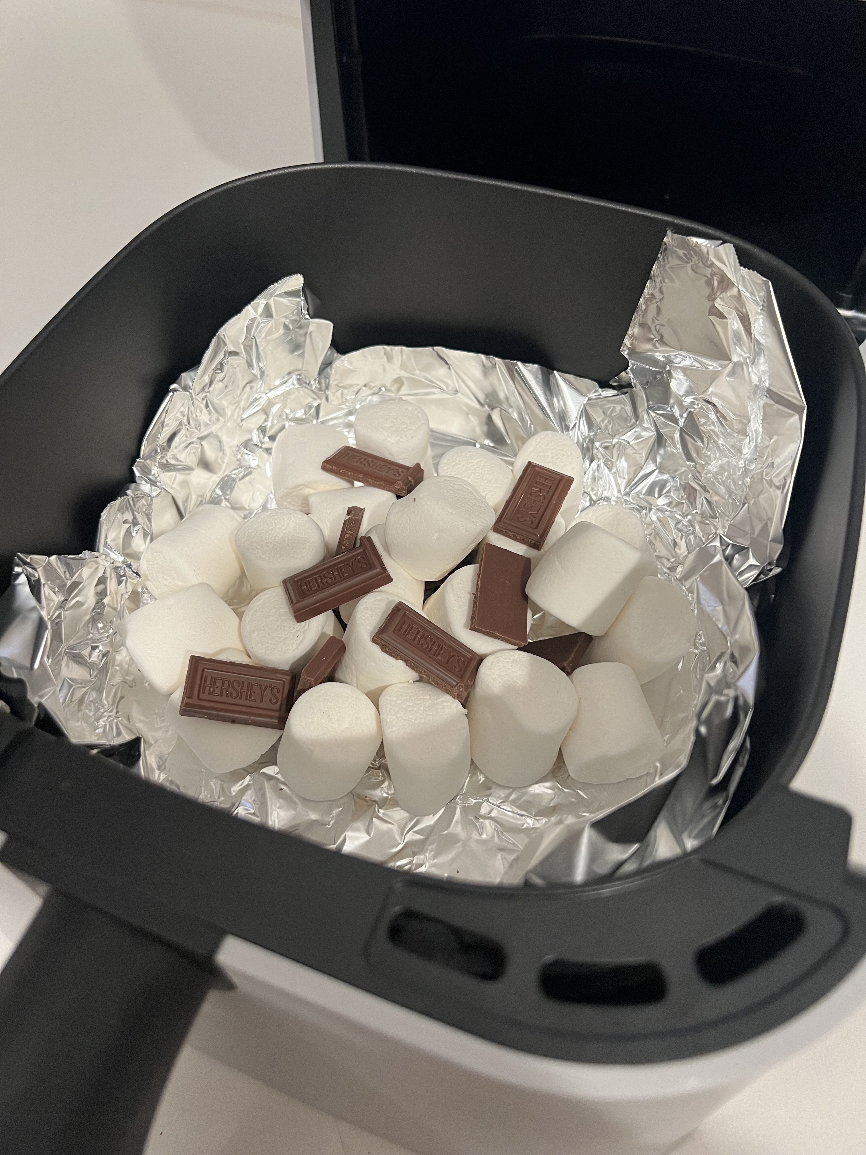 Marshmallows and Hershey's chocolate pieces on aluminum foil inside an air fryer, ready to be melted and combined