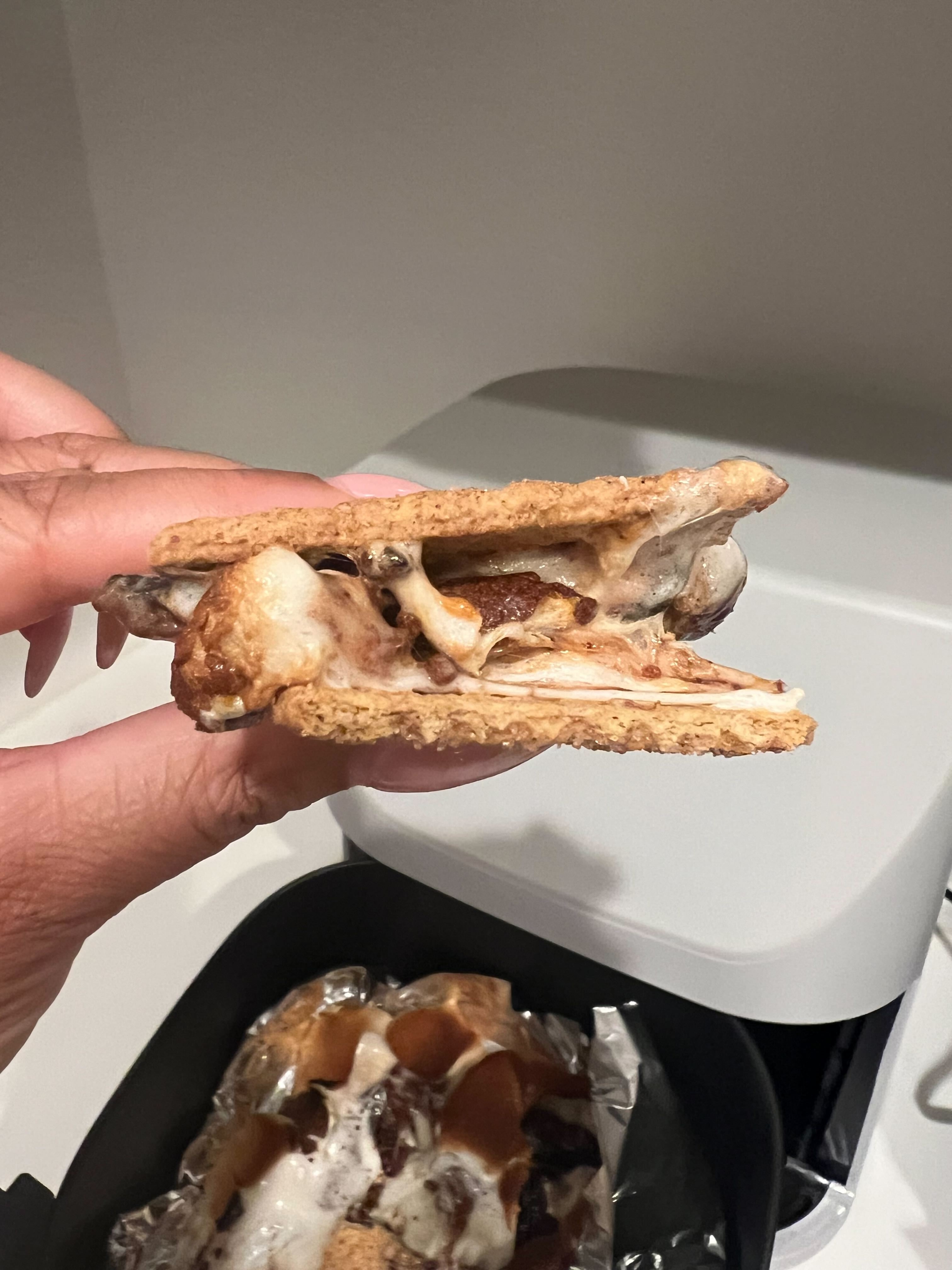 Close-up of a hand holding a gooey, freshly made s'more with melted marshmallow and chocolate. Another s'more is in an air fryer below