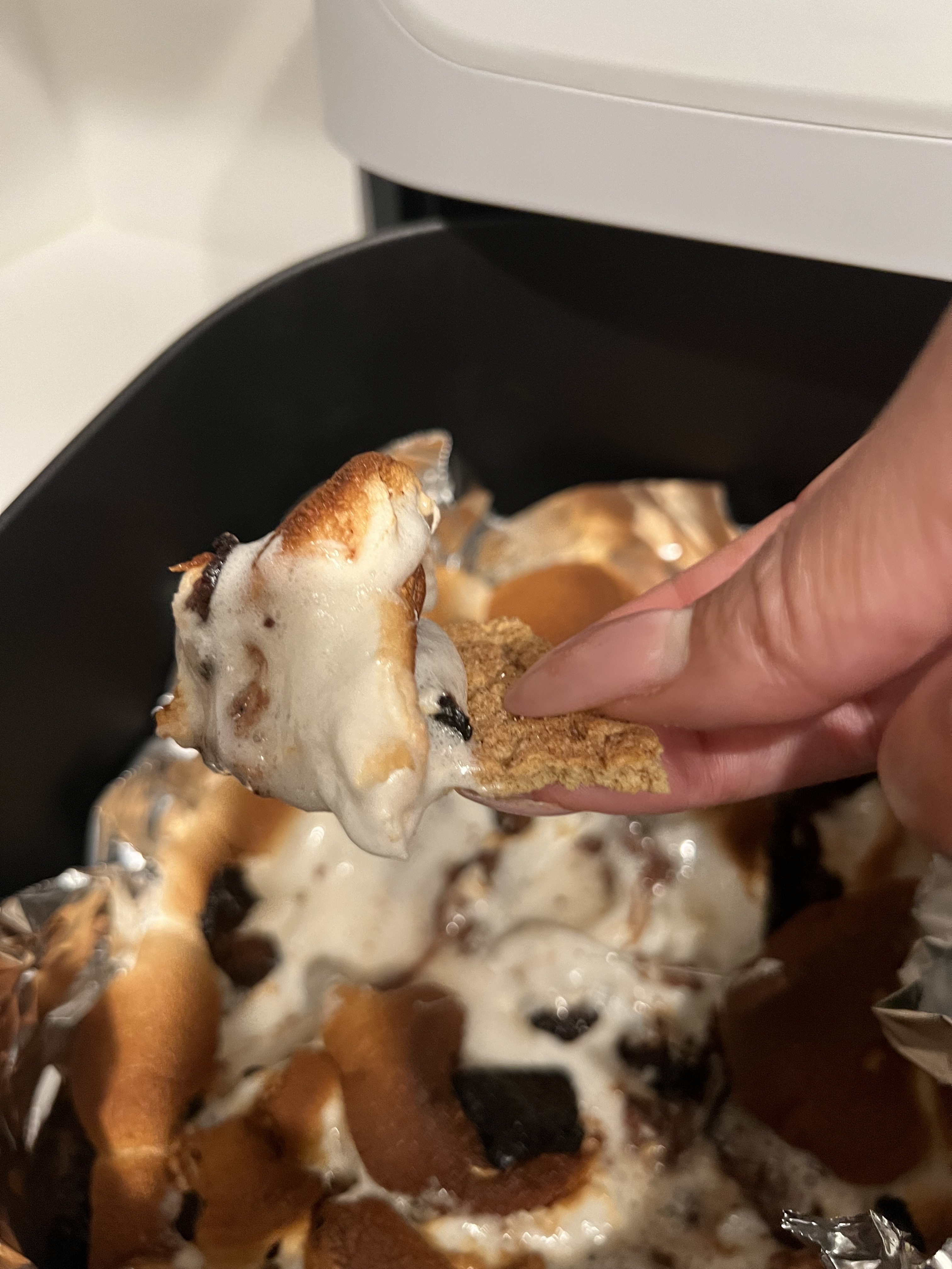A hand holds a piece of graham cracker dipped in melted marshmallows and chocolate from a S'mores dip in a foil-lined pan