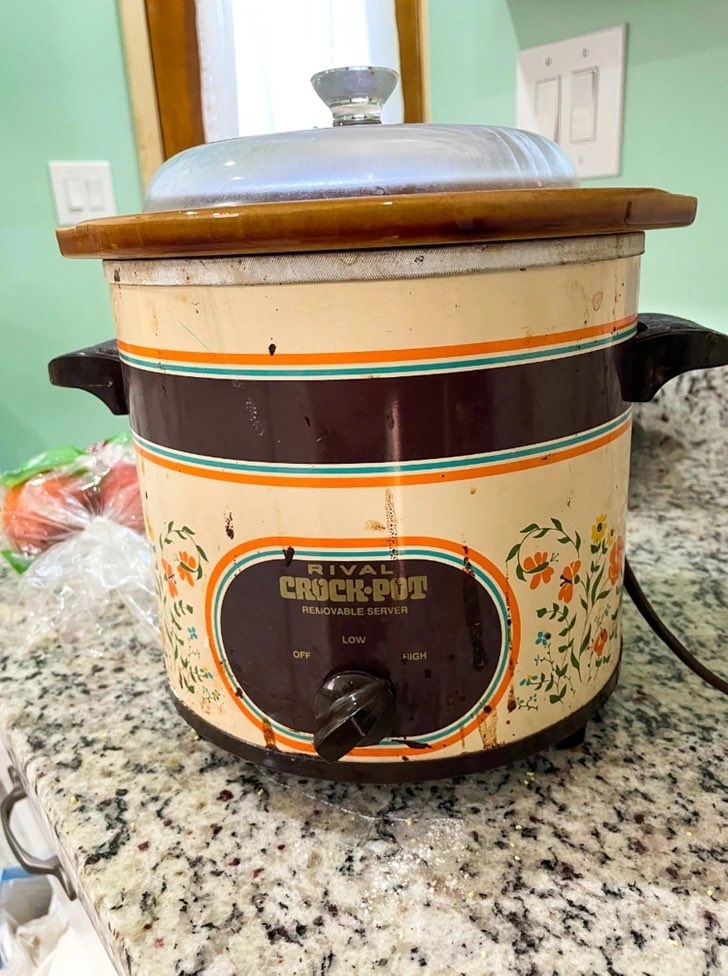 A Rival Crock-Pot slow cooker with colorful floral designs sits on a kitchen counter, with a glass lid on top and control dial set to "Low"