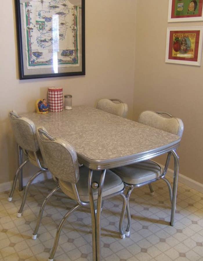 A retro dining room setup with a Formica table and four chairs, vintage wall art, and a salt and pepper shaker set on the table