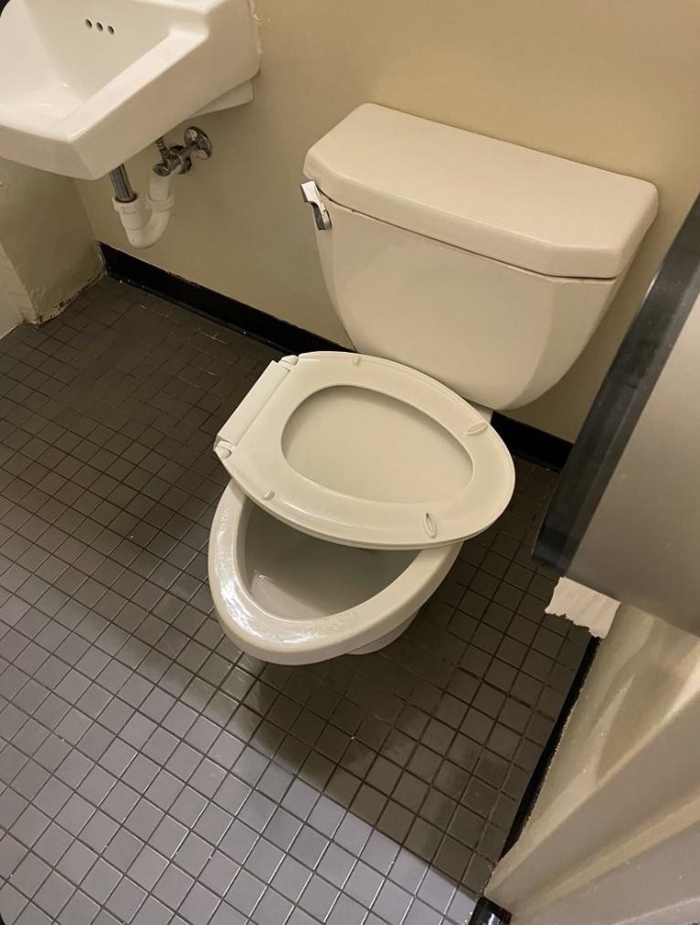 A toilet with the seat installed incorrectly, making it unusable, next to a small wall-mounted sink in a tiled bathroom