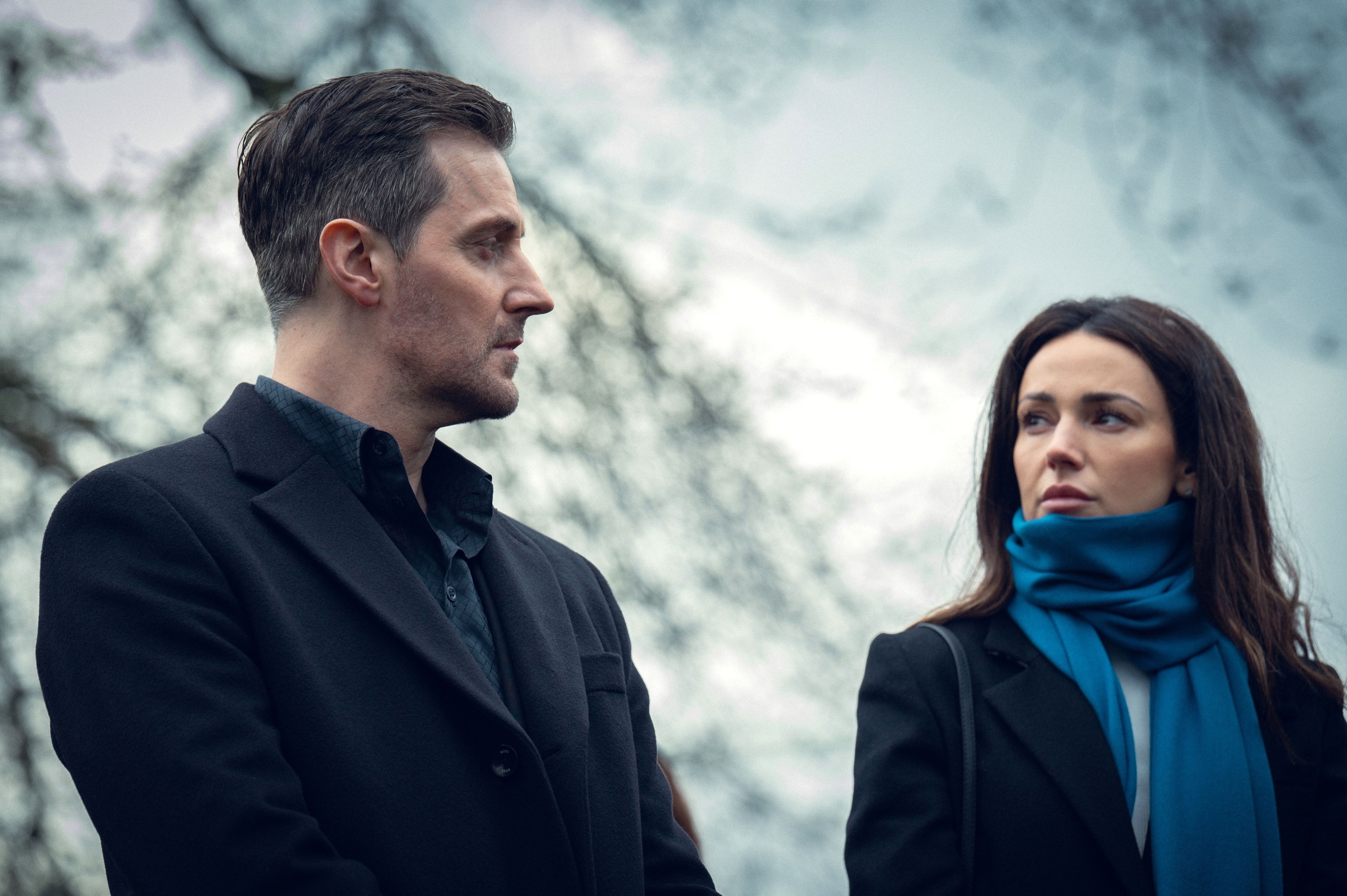 Richard Armitage and Michelle Keegan stand outdoors, facing each other seriously, both dressed in dark coats with Michelle wearing a blue scarf