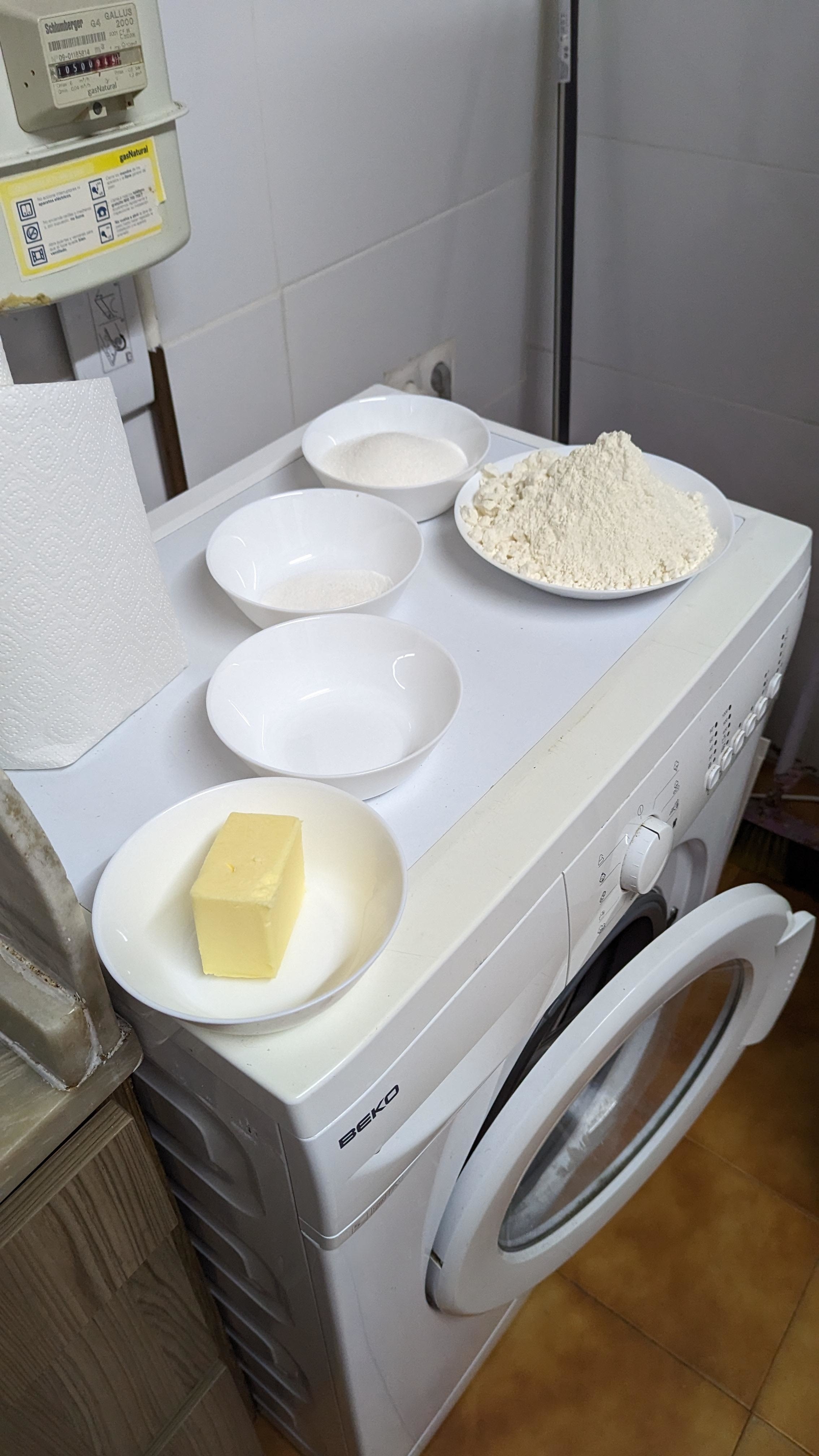 Four bowls: one with flour, one with sugar, one with salt, one with a stick of butter, are placed on a washing machine. A roll of paper towel is next to the bowls