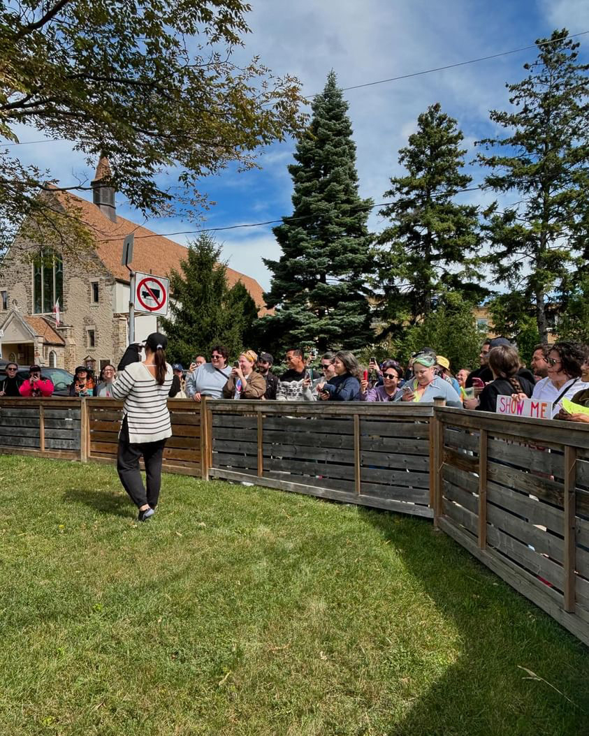 People standing behind a wooden barrier, possibly at an event or gathering, surrounding a grassy area near buildings and trees