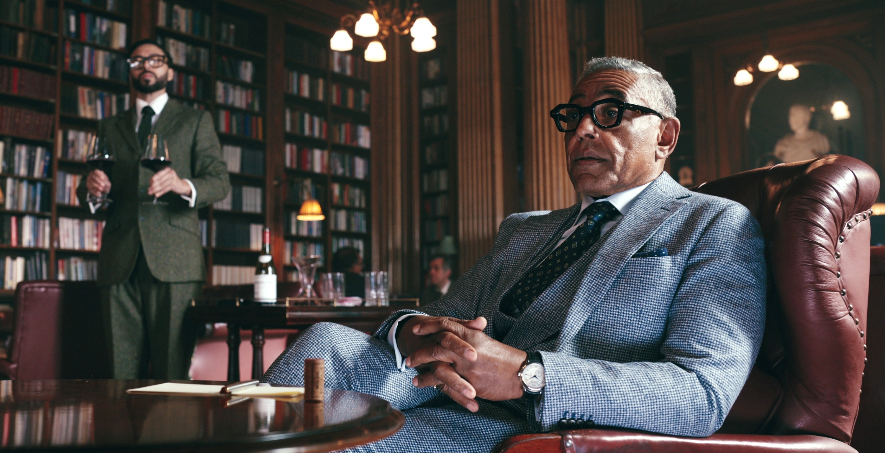 Giancarlo Esposito sits in a library in a stylish gray suit. A man in a suit stands in the background holding wine glasses