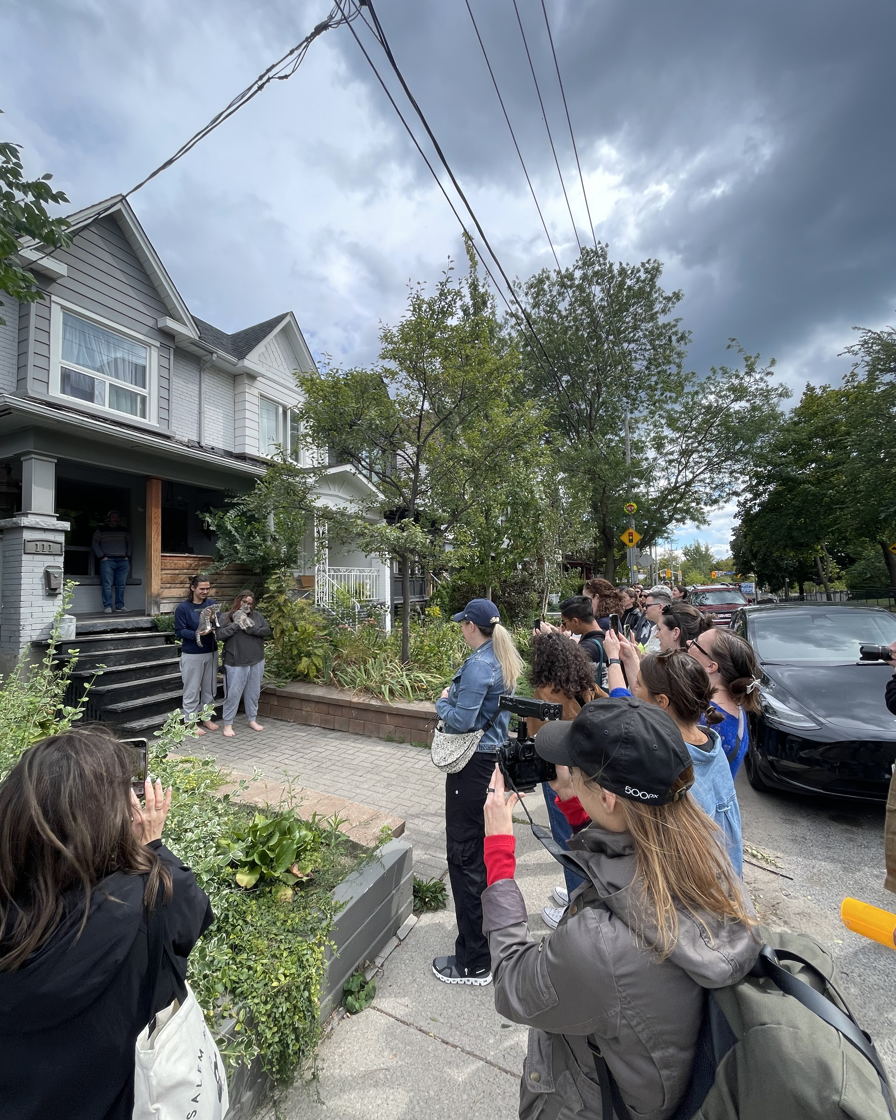 A group of people, some holding cameras, gather outside a residential home on a cloudy day, taking photos of a person standing on the porch