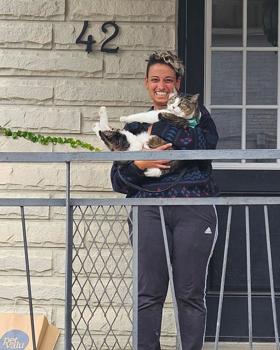 A person smiles while holding a large cat on a porch in front of a door numbered 42