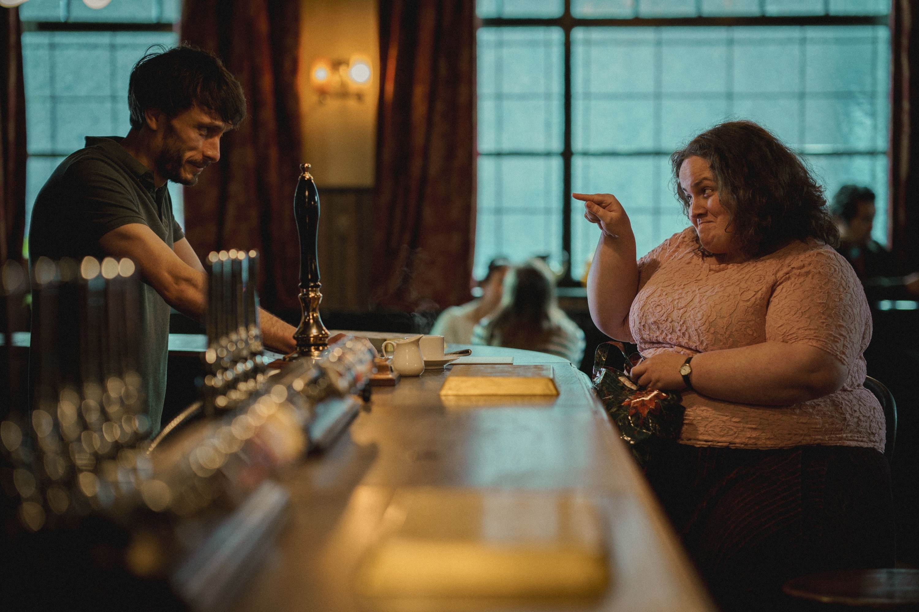 A man, sitting behind a bar counter, and a woman, leaning over the counter, engaged in conversation. The setting appears to be a pub or bar