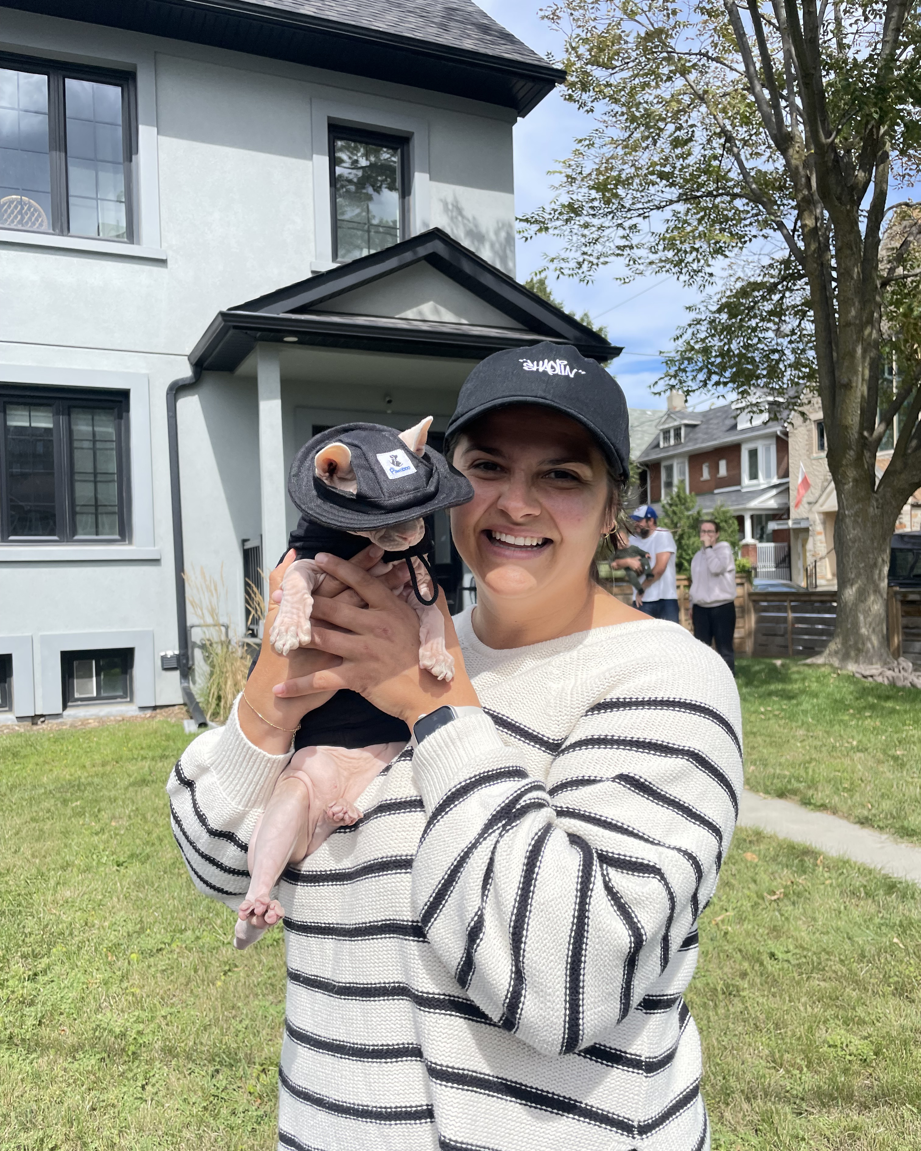 A smiling woman in a striped sweater holds a small dog wearing a hat and jacket outside a house