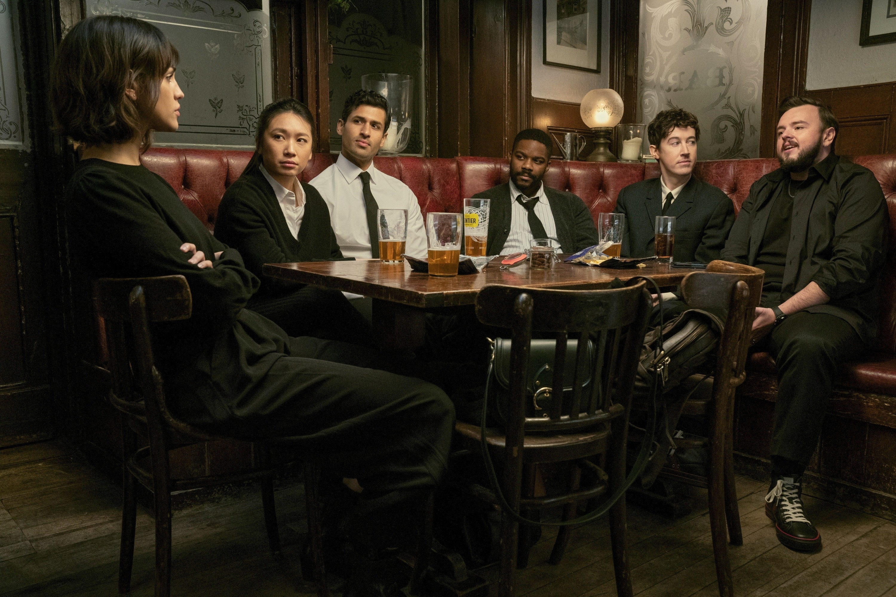 Group of six people, including Emma Appleton, Wunmi Mosaku, Nabhaan Rizwan, David Ajala, Edward Bluemel, and Tom Weston-Jones, sitting in a dimly lit pub