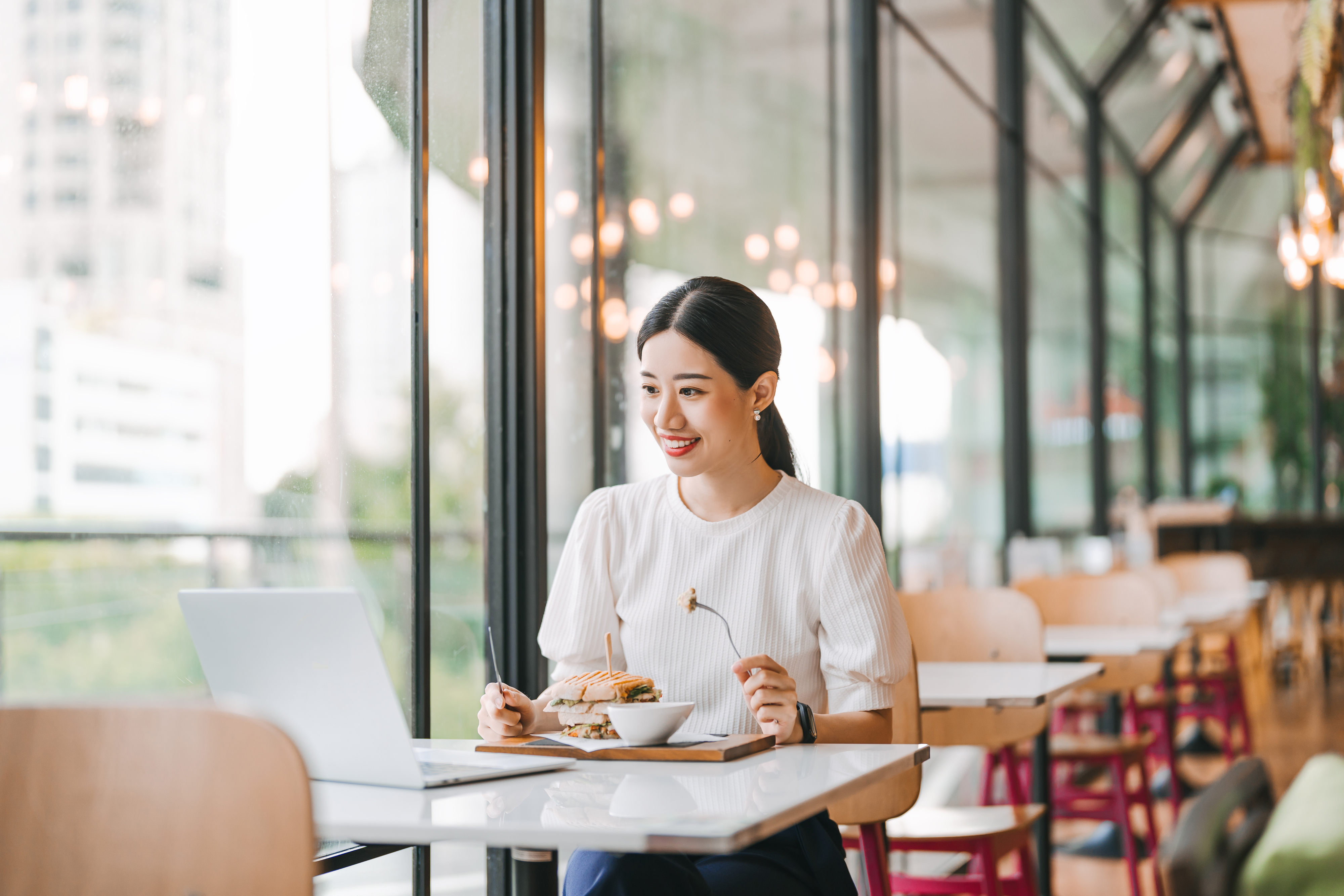 A woman sits at a table in a modern cafe. She is looking at a laptop and smiling, with a plate of food in front of her