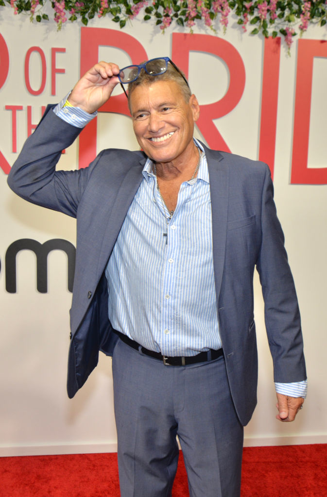 Steven Bauer on red carpet in a striped shirt and a blazer, smiling and holding up glasses