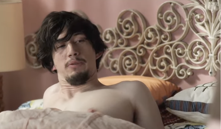 A shirtless man with messy hair lies in bed, appearing to have just woken up. An ornate headboard is visible behind him