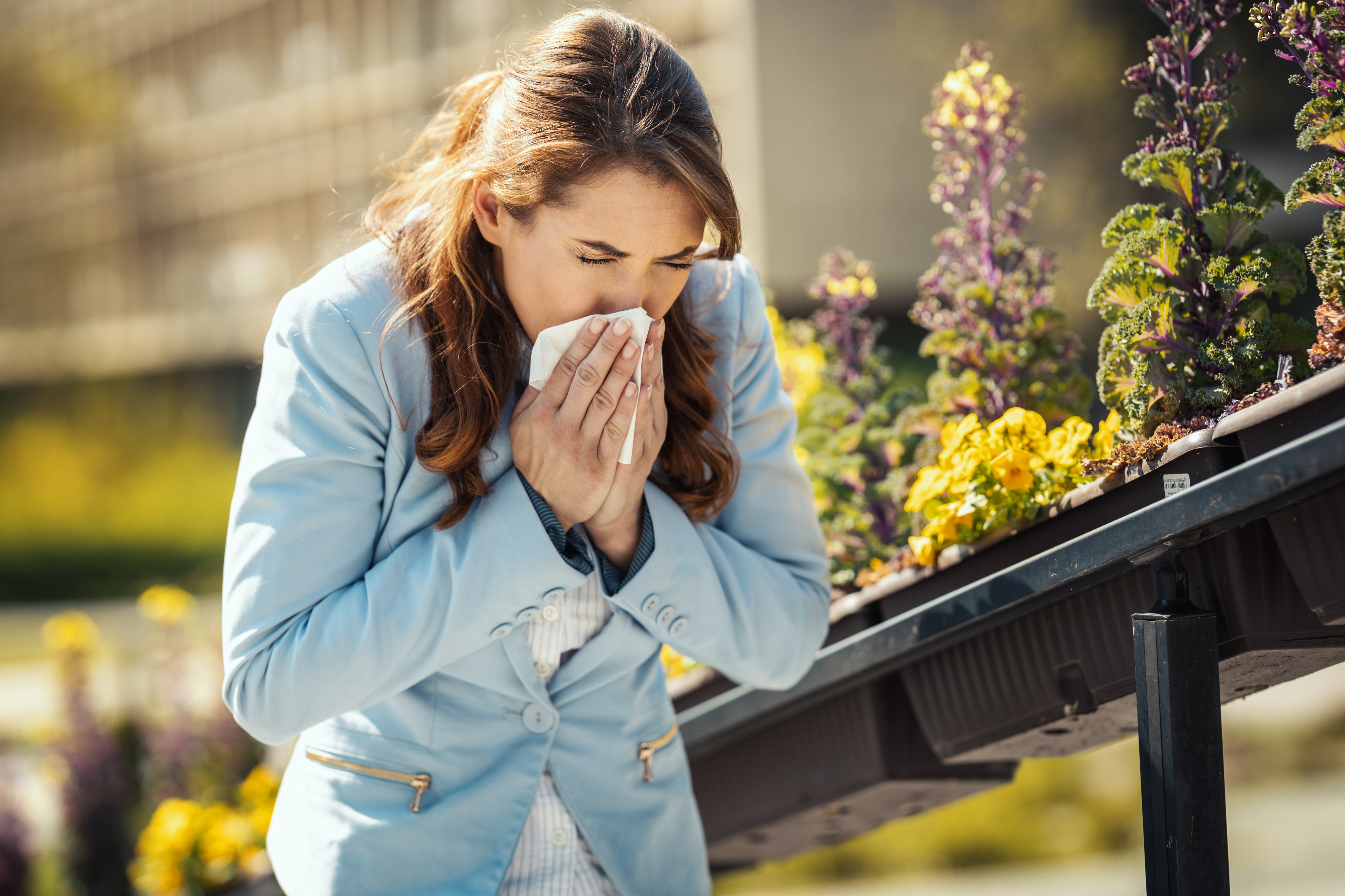 A woman in a blazer sneezes into a tissue while standing outdoors near blooming flowers