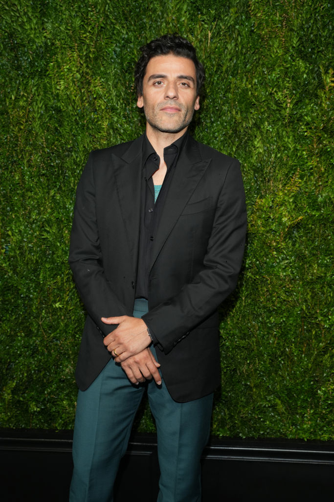 Oscar Isaac in a blazer and pants at an event, standing in front of a foliage background