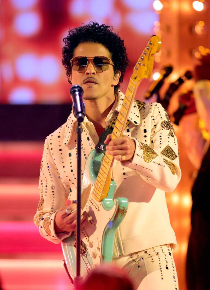 Bruno Mars performs on stage, playing a guitar, wearing a studded suit and sunglasses