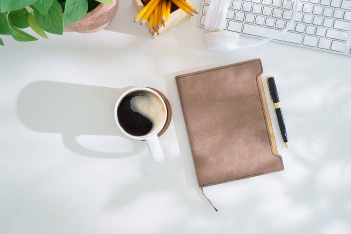 Journal, pen, coffee cup, keyboard, headphones, and pencils on a desk next to greenery