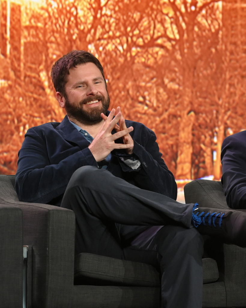 James Roday Rodriguez sits on a panel in casual business attire with a beard and a smile, hands clasped, legs crossed