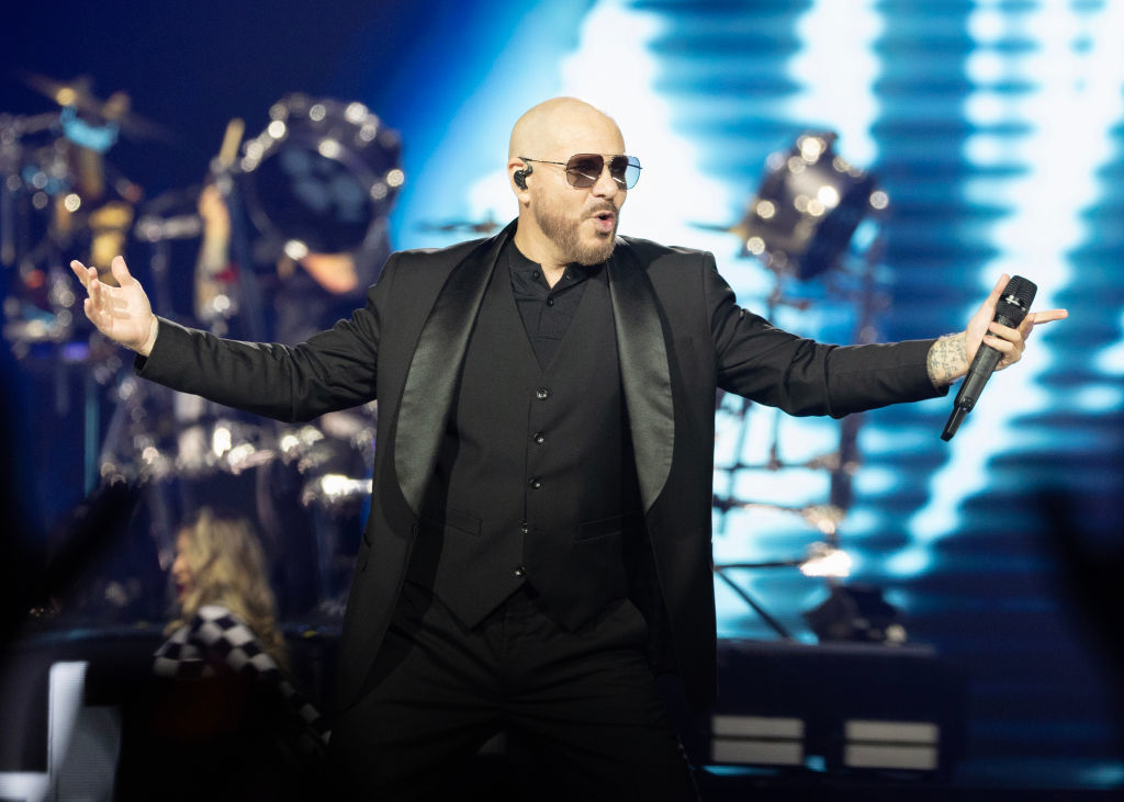 Pitbull performs onstage in a suit, holding a microphone, with a drummer in the background