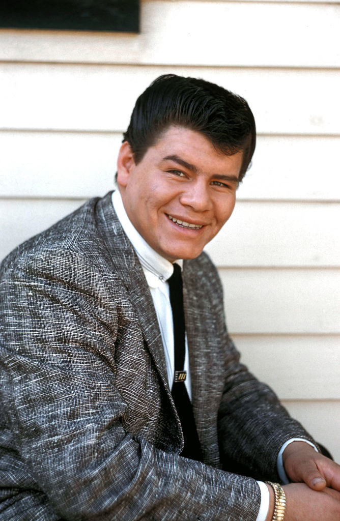 Ritchie Valens in a suit and tie smiles in a seated pose