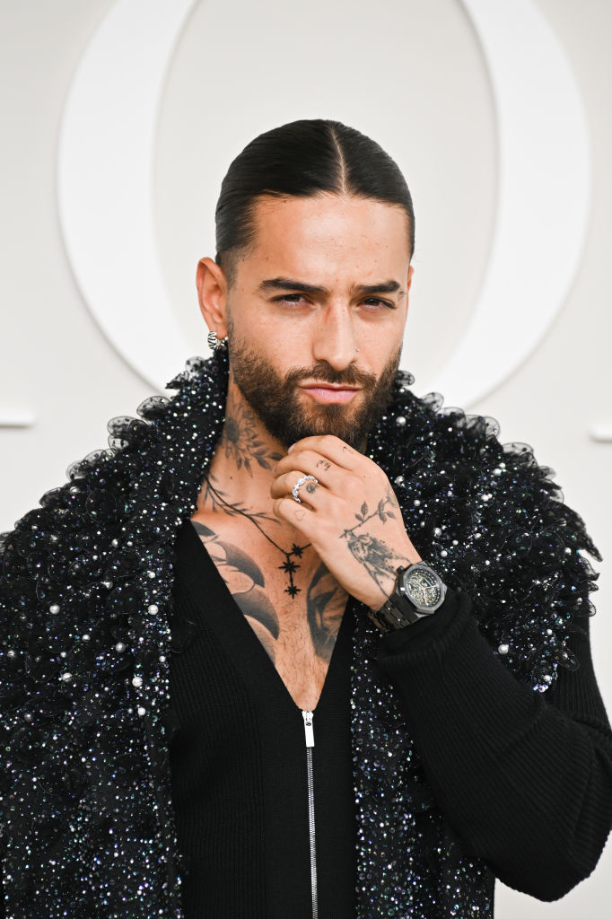 Maluma with slicked-back hair, beard, tattooed chest, wearing a shimmery, textured jacket and rings, posing thoughtfully with hand on chin