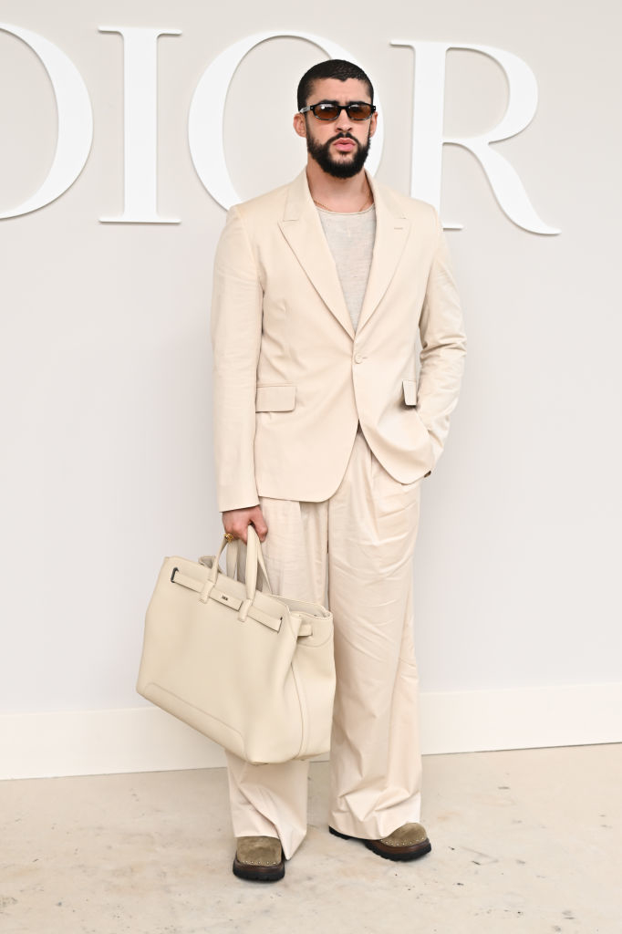 Bad Bunny in a light-colored suit, carrying a large bag, and wearing sunglasses, standing in front of a Dior backdrop
