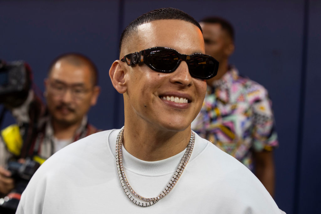 Daddy Yankee is smiling, wearing sunglasses, a t-shirt, and layered necklaces at a public event. Photographers are visible in the background