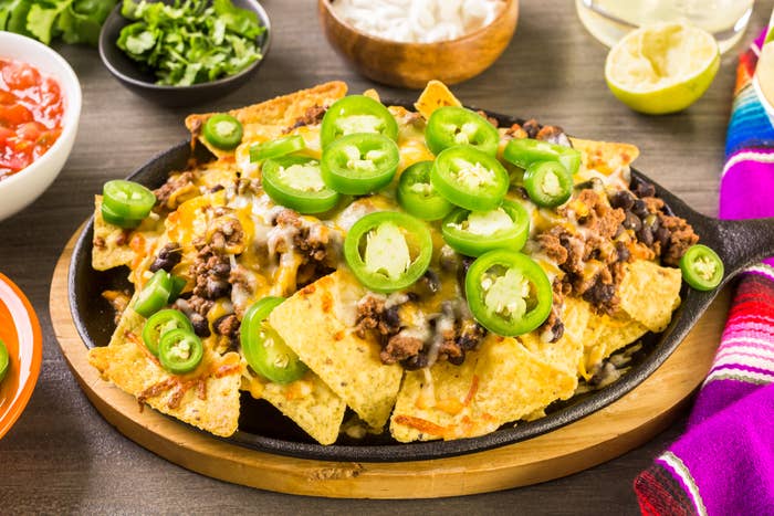 A plate of nachos topped with melted cheese, ground beef, and sliced jalapeños, placed on a wooden serving tray surrounded by condiments in bowls