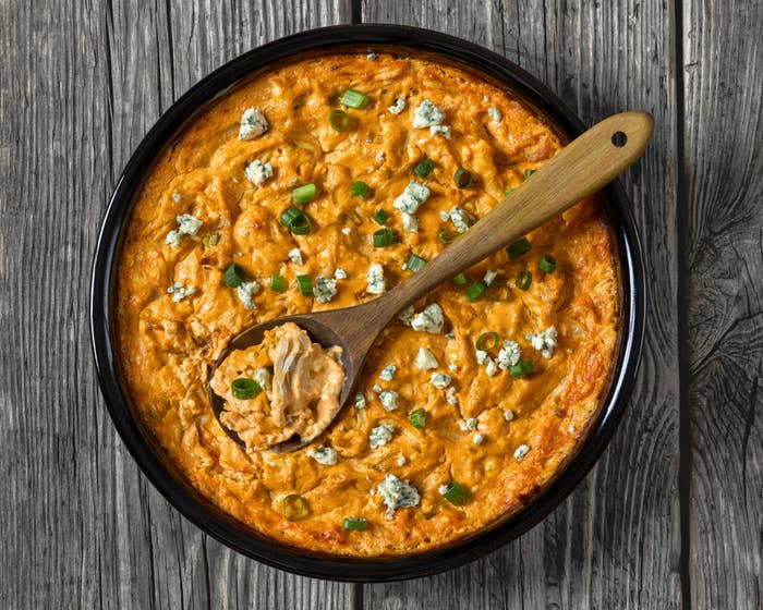 A round dish of buffalo chicken dip garnished with green onions and blue cheese crumbles. A wooden spoon is placed in the dip