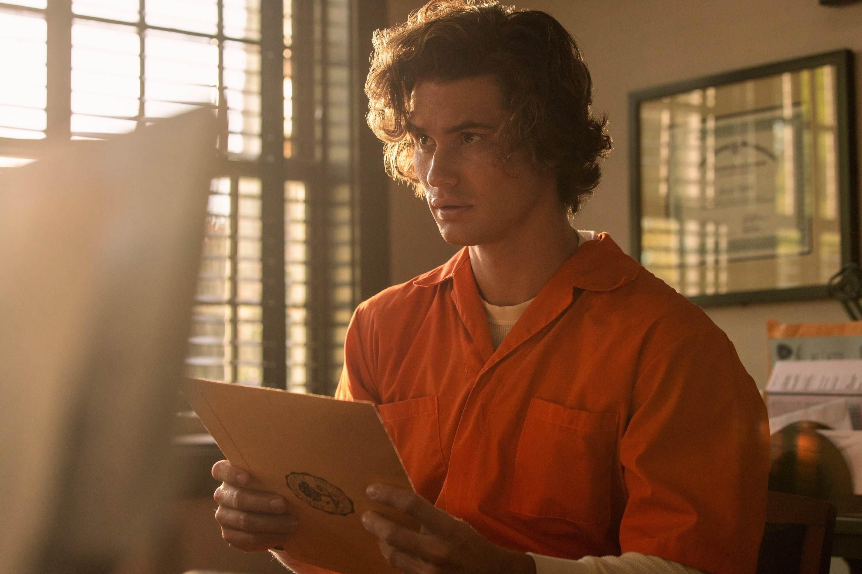 Chase Stokes wearing an orange jumpsuit, holding an envelope in "Outer Banks"