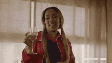 Amanda Seales from Insecure raising a wine glass and smiling, wearing a casual sporty jacket and pigtails