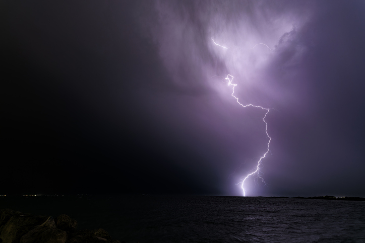Lightning bolt striking the horizon under a night sky, captured over a body of water, creating a dramatic and intense atmospheric scene. No people are present