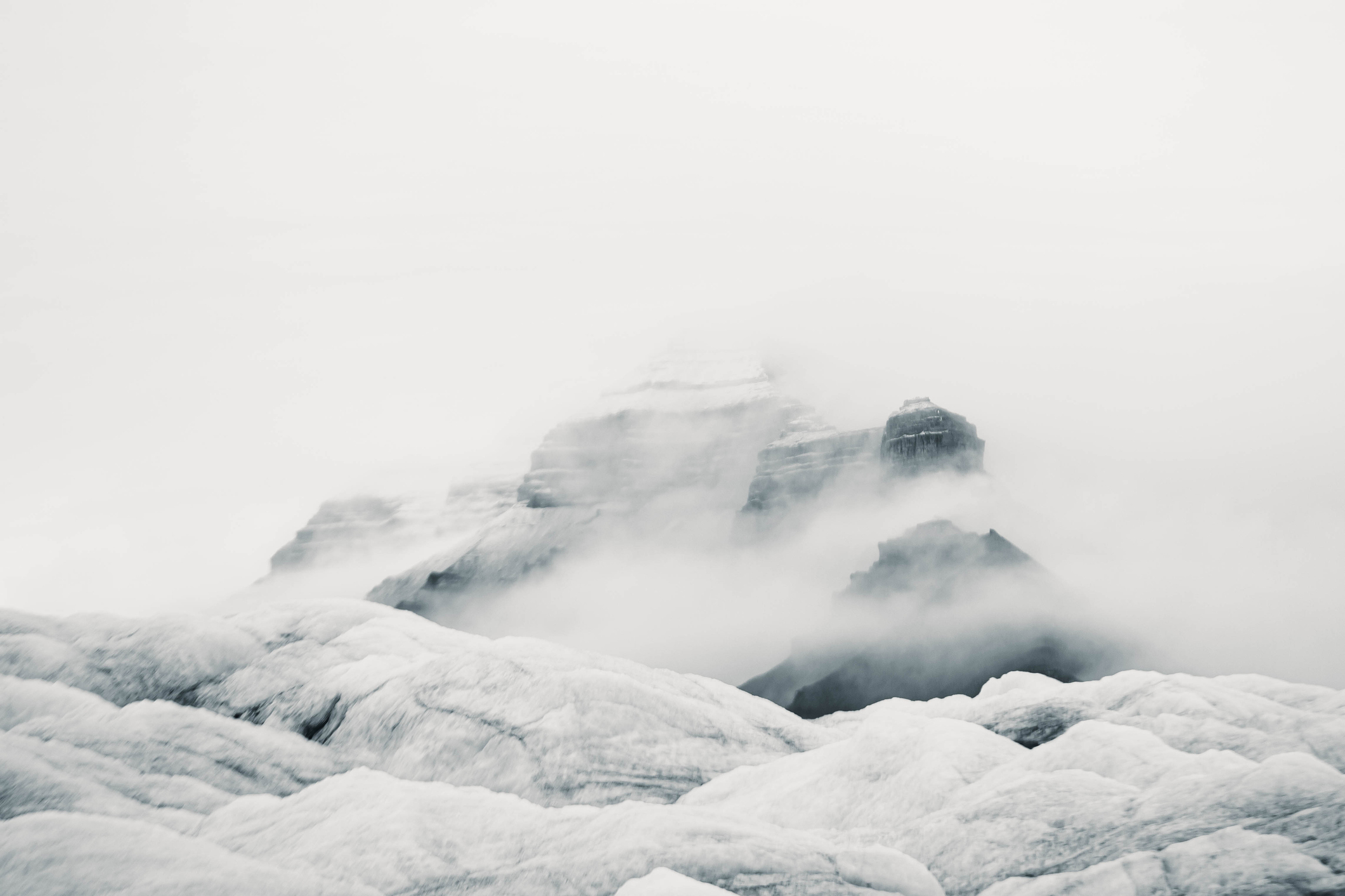 A misty mountain peak partially obscured by clouds, with rugged, snow-covered terrain in the foreground