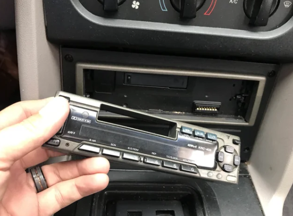 A hand is holding a detached car stereo faceplate in front of its empty slot on the car dashboard. The dashboard shows climate control knobs above