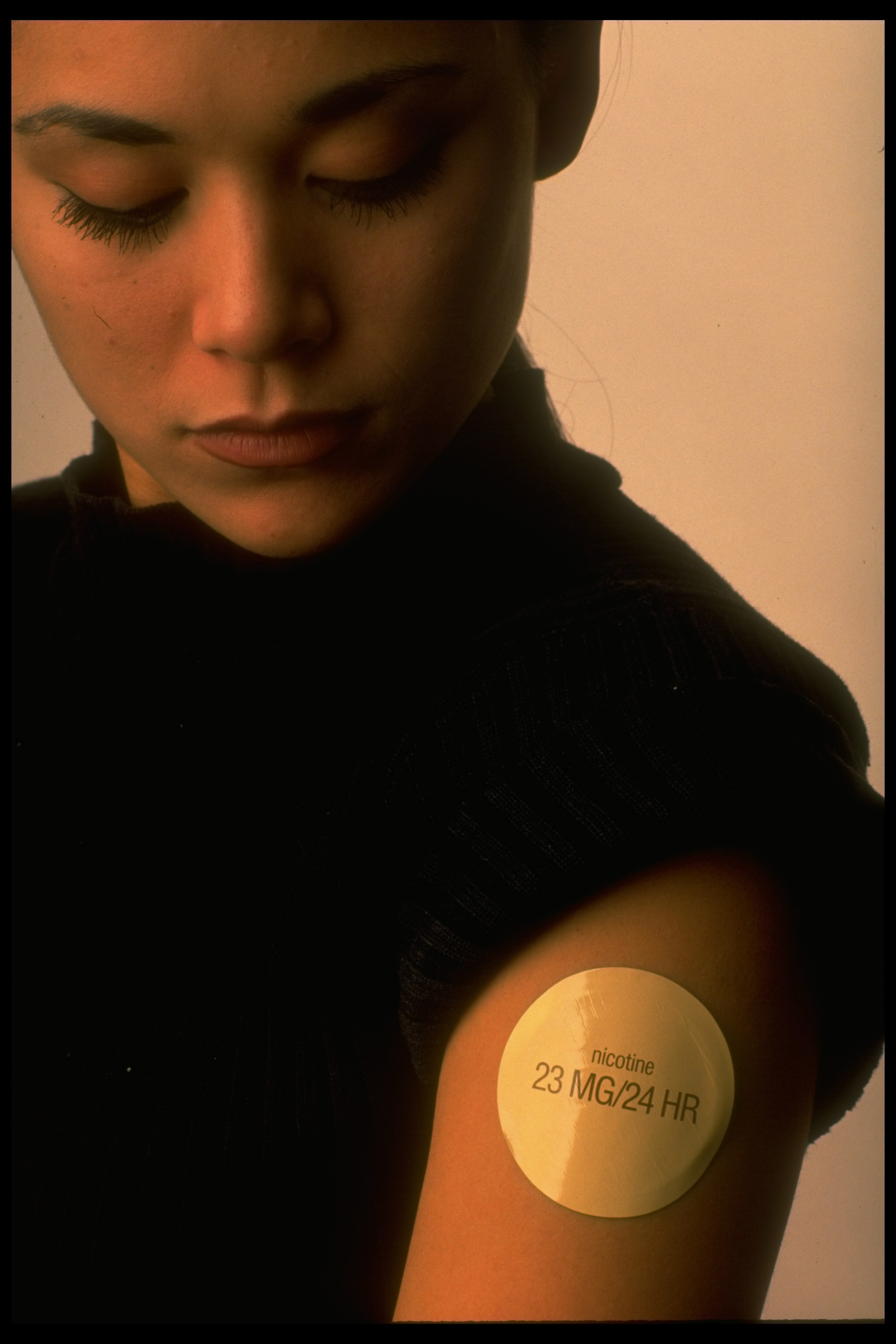A woman looks down at a nicotine patch on her upper arm that reads "nicotine 23 MG/24 HR."