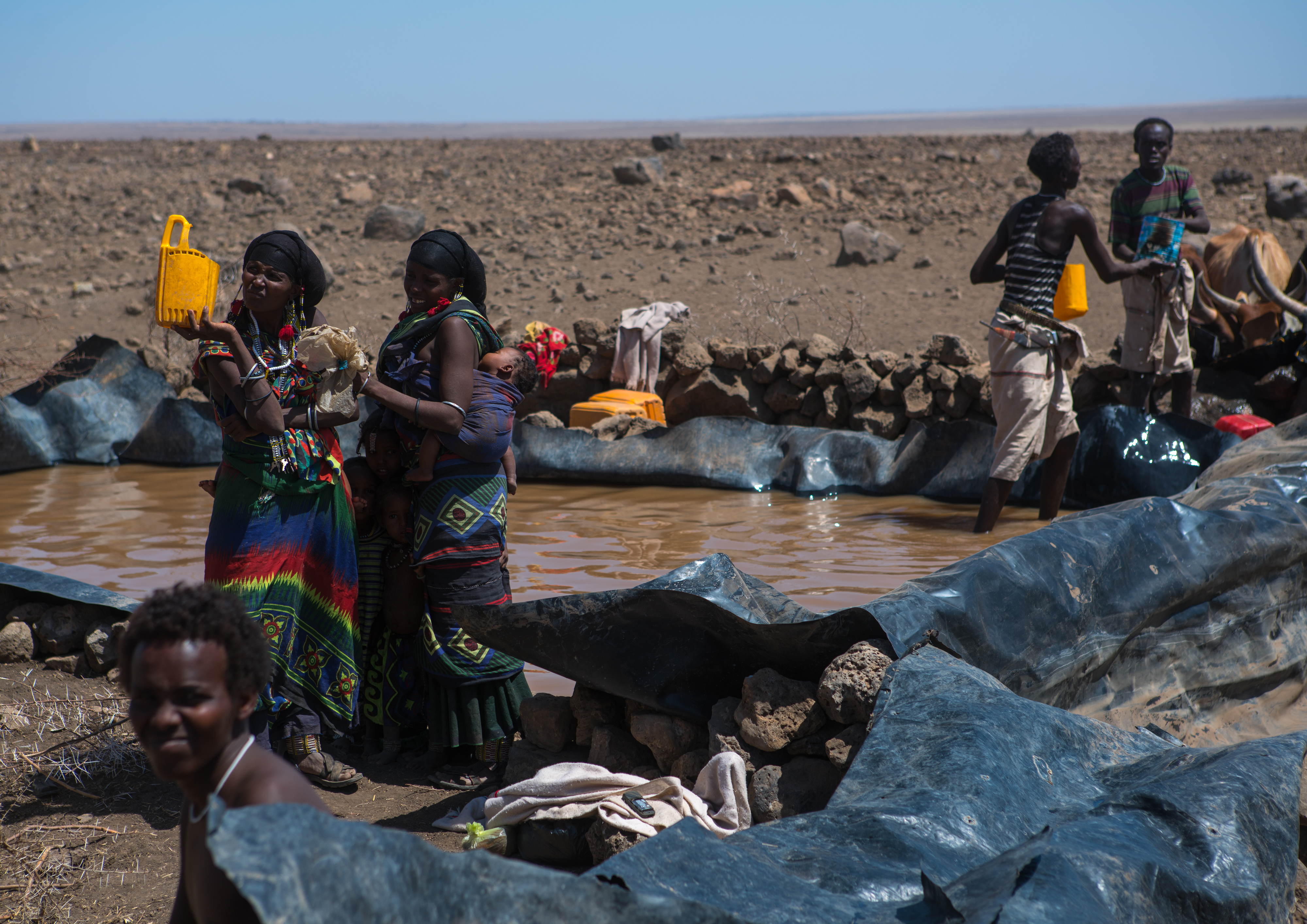 People gather near a makeshift pond in a dry, arid landscape. They fill containers with water, possibly for drinking or daily use