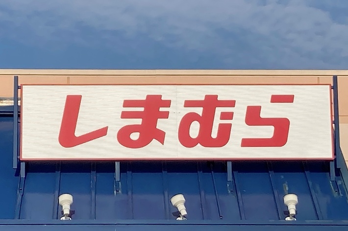 The image shows a storefront sign with Japanese text that reads “しまむら.” The store facade has a blue awning and a handicap parking sign