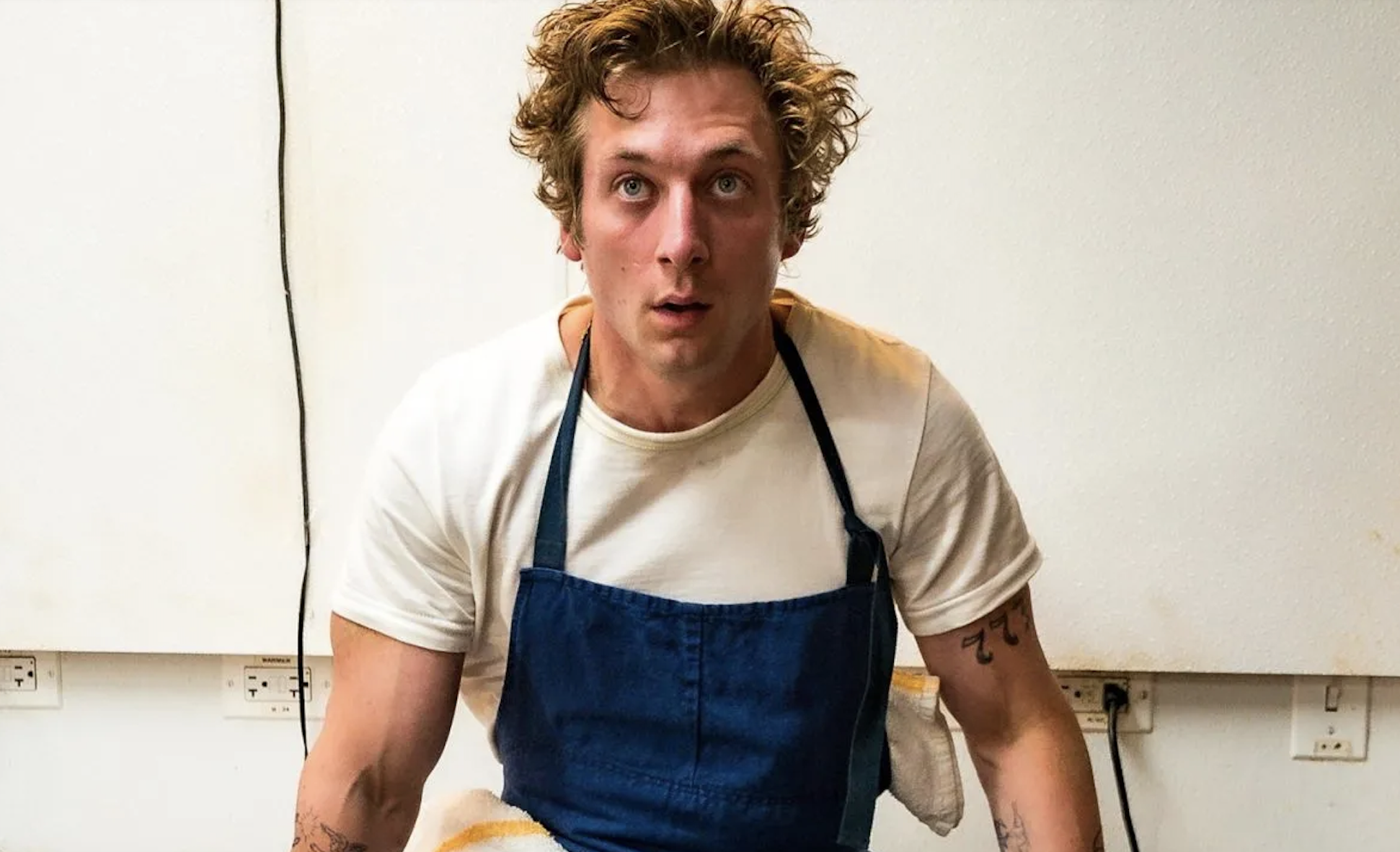 Jeremy Allen White wears a white T-shirt and blue apron in a kitchen setting, looking intently ahead, with visible tattoos on his arms