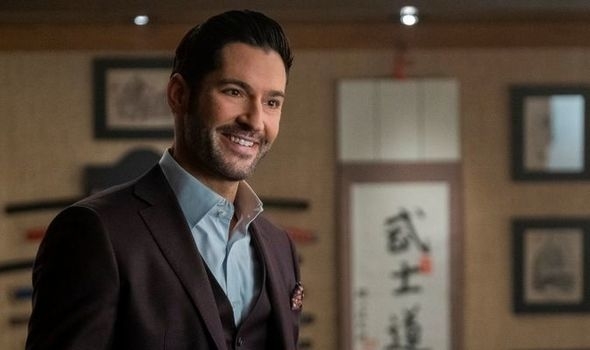Tom Ellis, smiling, in a suit with an open-collared shirt, appears in a room with Asian-inspired decor, including framed calligraphy and art