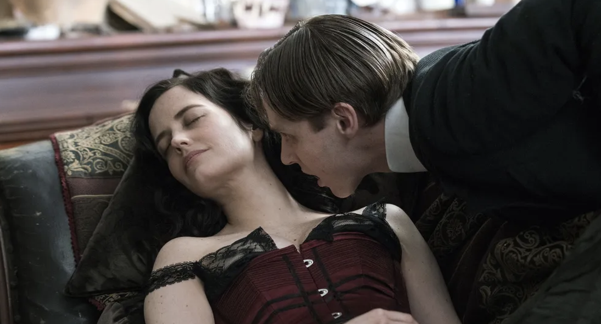 Eva Green lies on a couch wearing a dark, Victorian-style dress. A man is leaning over and looking at her intently