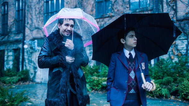 Two individuals, one holding a clear umbrella with a pink rim, the other holding a black umbrella, stand outside near a building. The man on the left is smiling, while the boy on the right looks serious