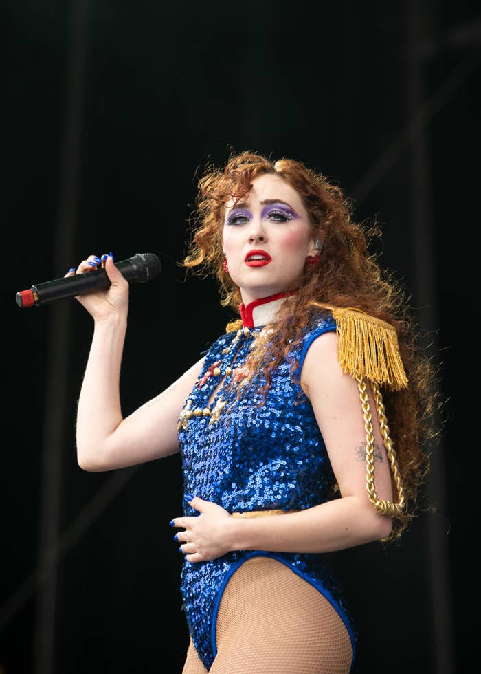 A woman with curly hair performs on stage, wearing a sequined bodysuit with gold fringe epaulets and holding a microphone