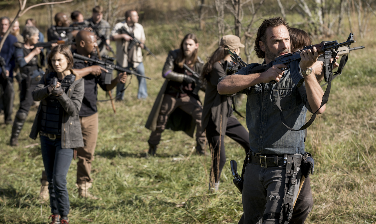 A group of people, including Rick Grimes, Daryl Dixon, and Michonne from "The Walking Dead," stand armed and ready in an outdoor, grassy area
