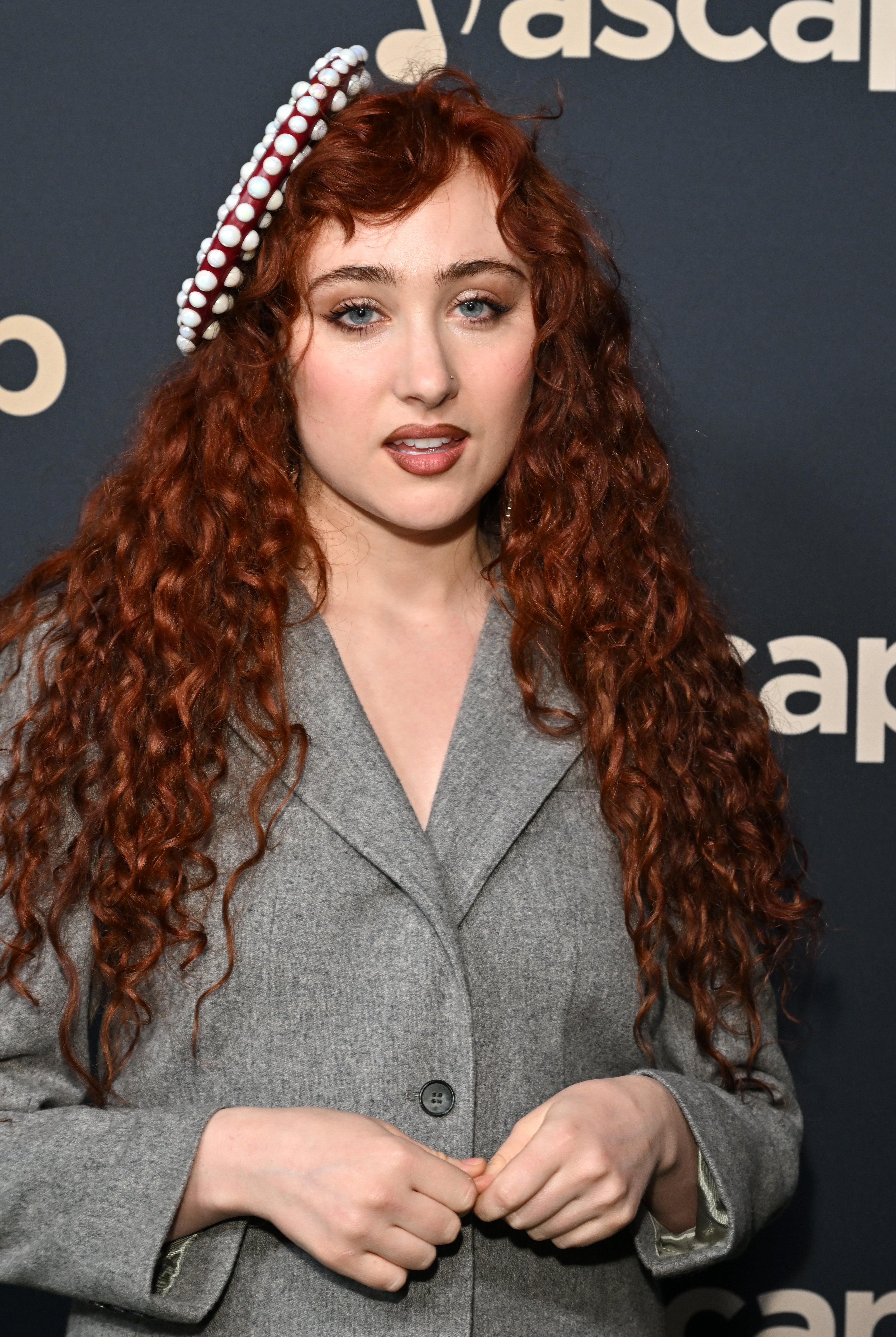 A person with long curly hair, wearing a grey outfit and a headpiece with decorative accents, poses on the red carpet at an ASCAP event backdrop