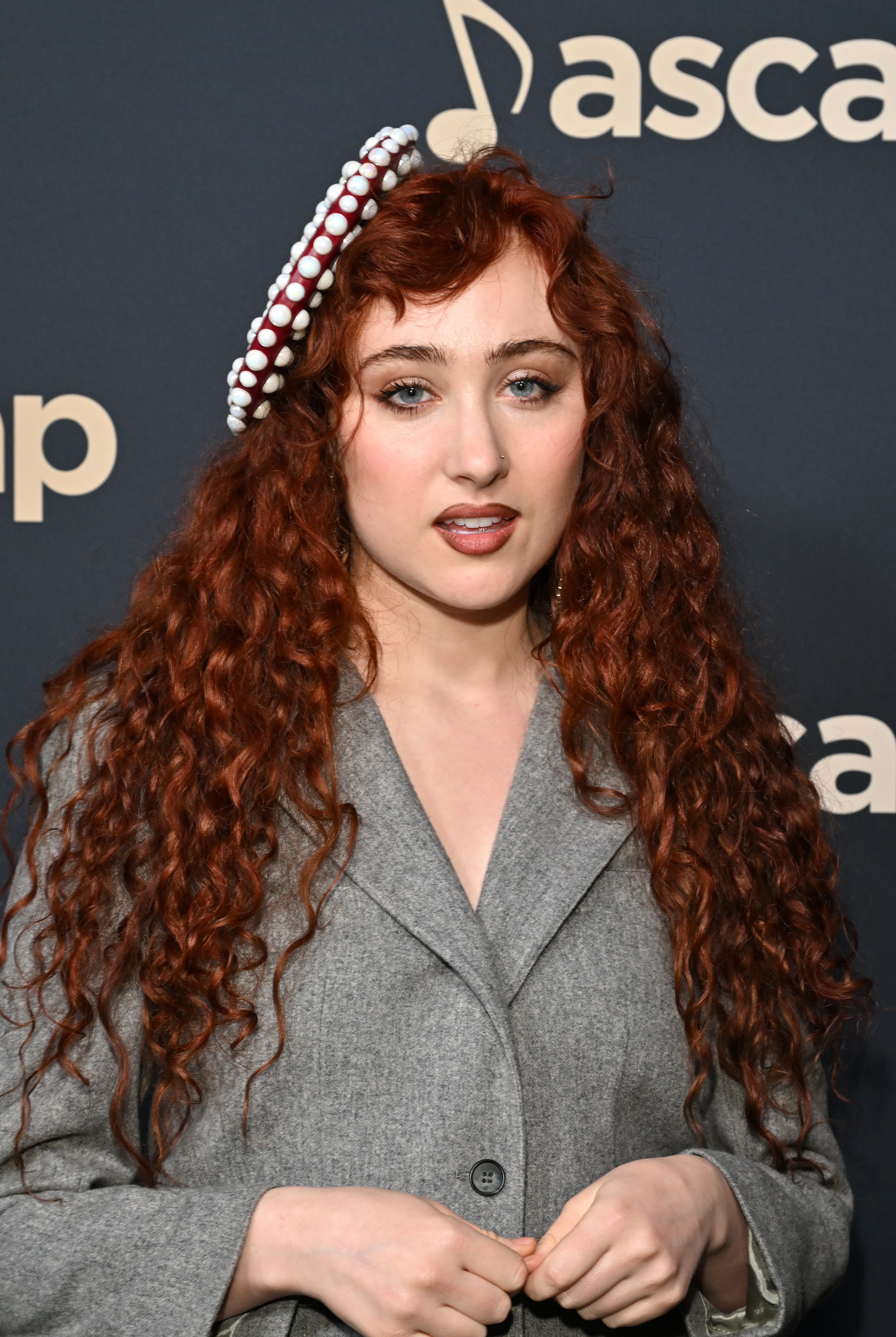 A person with long curly hair, wearing a grey outfit and a headpiece with decorative accents, poses on the red carpet at an ASCAP event backdrop