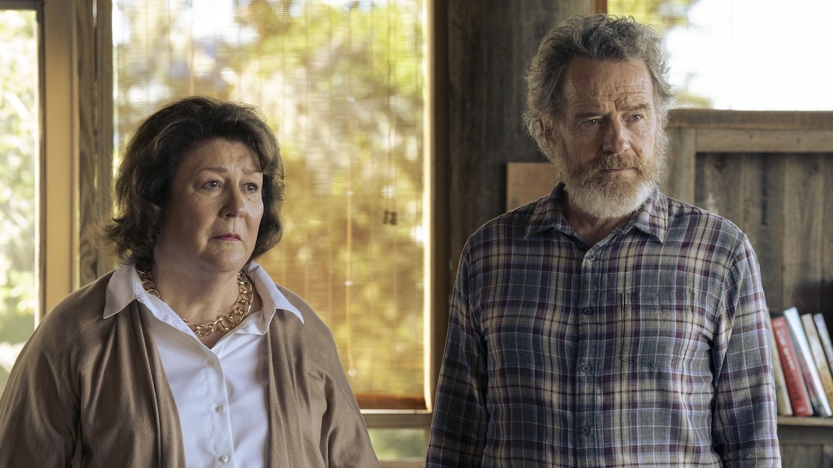 Bryan Cranston and Margo Martindale are standing side by side in a rustic setting. Bryan wears a plaid shirt, while Margo wears a blouse with a sweater and a necklace