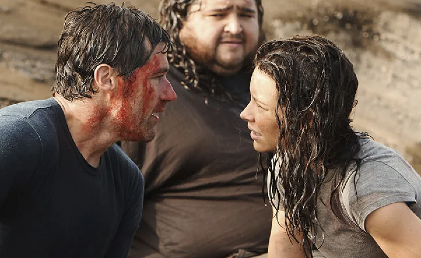 Matthew Fox, Jorge Garcia, and Evangeline Lilly are standing on a beach. Matthew Fox has blood on his face, and they appear intense, as if engaged in a serious conversation
