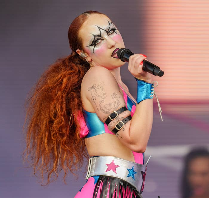 Chappell Roan performs on stage wearing a vibrant costume with star motifs and bold makeup. She holds a microphone and poses with her head turned to the side